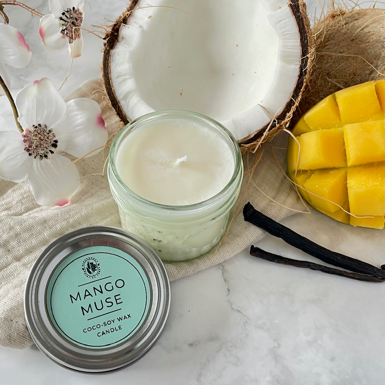 Mango Muse candle in a jar with coconut, mango, and vanilla beans on a marble surface.